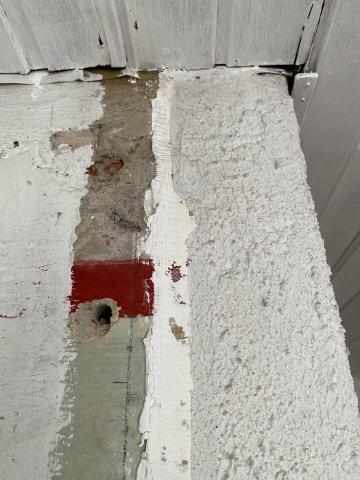 Skalad, flagnad vitmålning på vägg och golv med exponerad röd yta och hål. Behov av underhåll eller reparation.