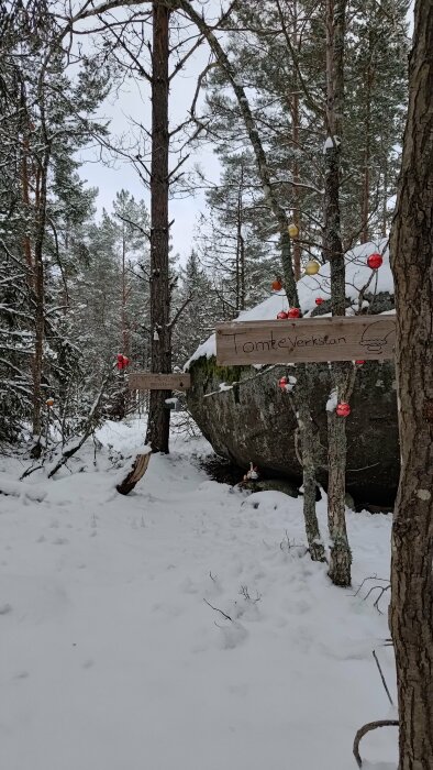 Vinterskog med skylt "Tomteverkstan", julpynt, stor sten och snö.