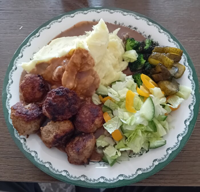 En måltid med köttbullar, potatismos, brun sås, grönsaker och sallad på ett mönstrat fat.