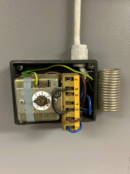 Öppen termostat med inställningsratt, ledningar, temperatursensor och tejp, monterad på vägg.