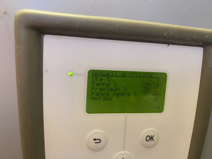 Digital display visar aktuell driftinformation för värmeanläggning med temperaturvärden; omgiven av damm och spindelnät.