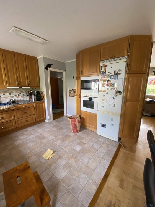 Kök med träskåp, vitvaror, kaklat golv, katt på dörrkarm, kylvägg med foton, påse på golvet.