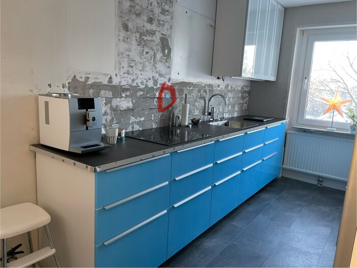 Kök med blå skåp, kaffemaskin, ej färdigställda väggar, fönster med julstjärna, grått golv.