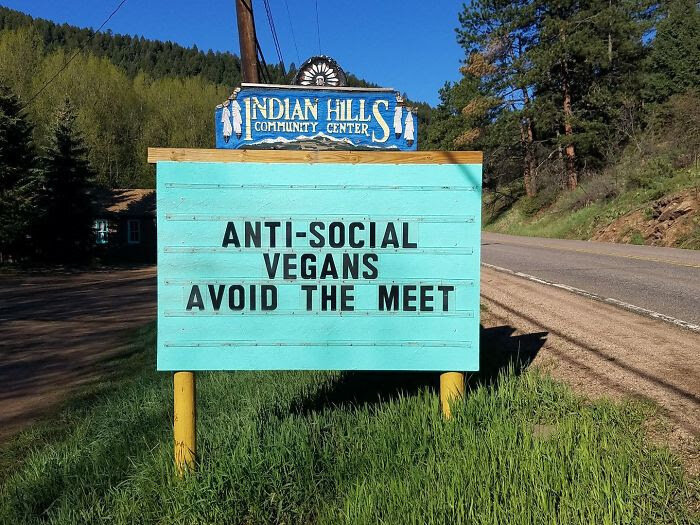 Turkos skylt med ordlek, "ANTI-SOCIAL VEGANS AVOID THE MEET", i naturen vid en väg.