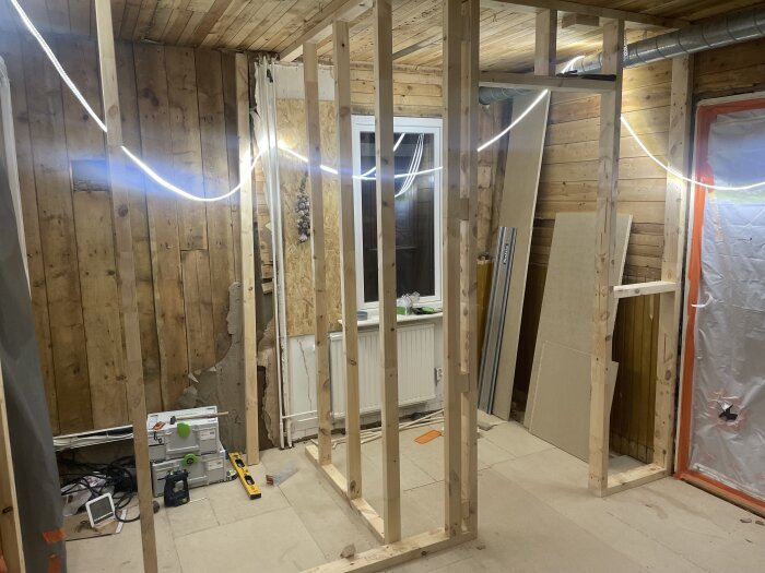 Renoveringsarbete med träreglar, isoleringsskivor, verktyg och byggmaterial i ett oavslutat rum.