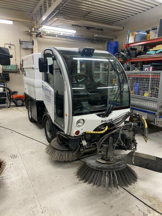 Vit sopbil med roterande borstar i ett garage, med diverse utrustning och hyllor i bakgrunden.