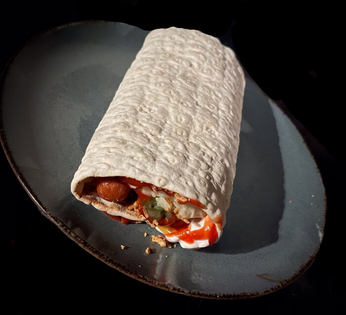 Korv i bröd med såser och grönsaker, rullad i tortillabröd på mörk tallrik.