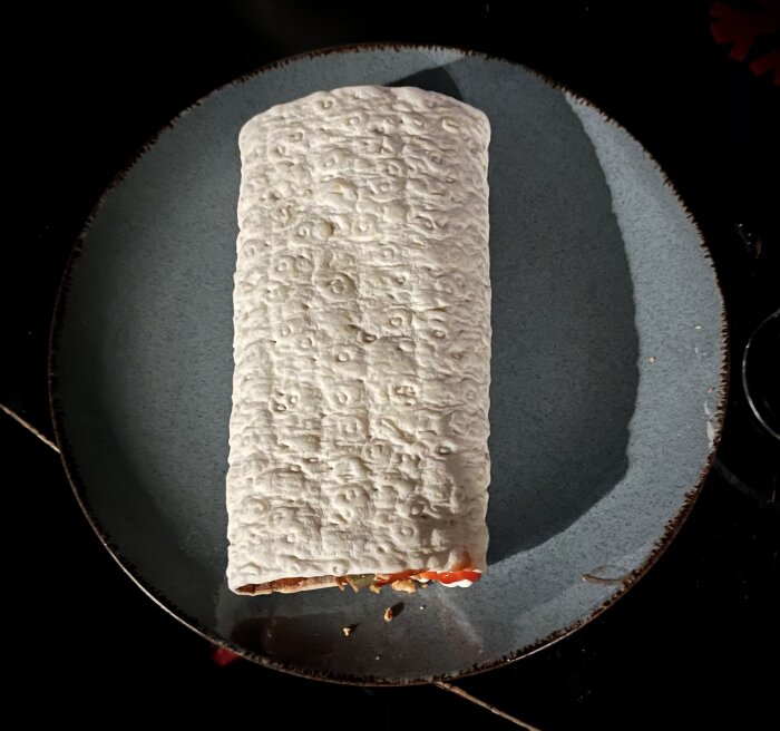 En rullad tortilla med fyllning på en mörk tallrik. Ljuset reflekterar på ytan. Snabbmat eller hemlagat.
