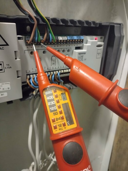 Multimeter mäter spänning på elcentral, visar cirka 240 volt, säkringar och anslutna kablar i bakgrunden.