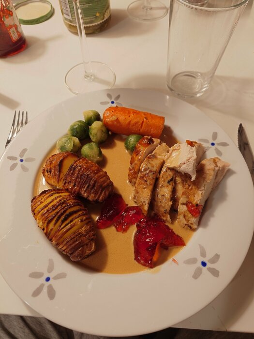 En tallrik med hasselbackspotatis, kyckling, brysselkål, morot och röd sås. Ett glas och bestick syns också.