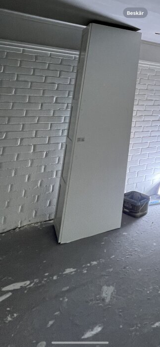 Ett stort, vitt skåp står lutat i ett garage vid en tegelvägg. Nedskräpat golv, grått.