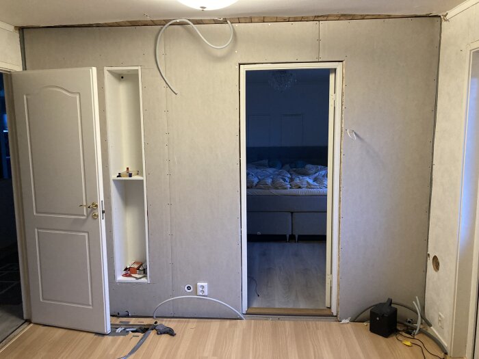 Renovering pågår: oavslutade gipsskivor, exponerade kablar, verktyg på golvet, synlig dörr till sovrum.