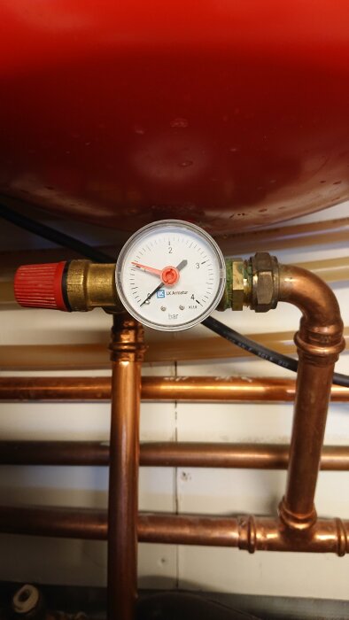 Manometer på kopparledning visar tryck, installerad under röd behållare, eventuellt del av värmesystem.