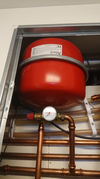 Röd expansionskärl med tryckmätare monterat över kopparledningar och rörstöd. VVS-installation, uppvärmningssystem.