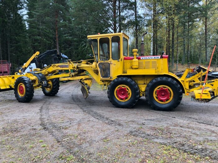 Gul Volvo BM grävmaskin på grus med skogsbakgrund.