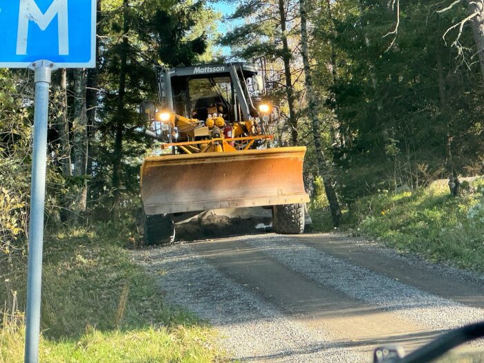 Gultonad hjullastare med skopa korsar grusväg vid blått M-vägmärke, omgärdad av skog i solljus.