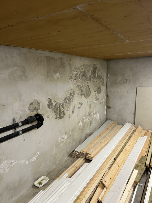 Källarrum med fukt- eller mögelskador på vägg, träreglar och vita lister på golvet.