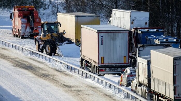 Lastbilar i kö i vinterväder, snöröjning pågår med hjälp av traktor och plogbil.