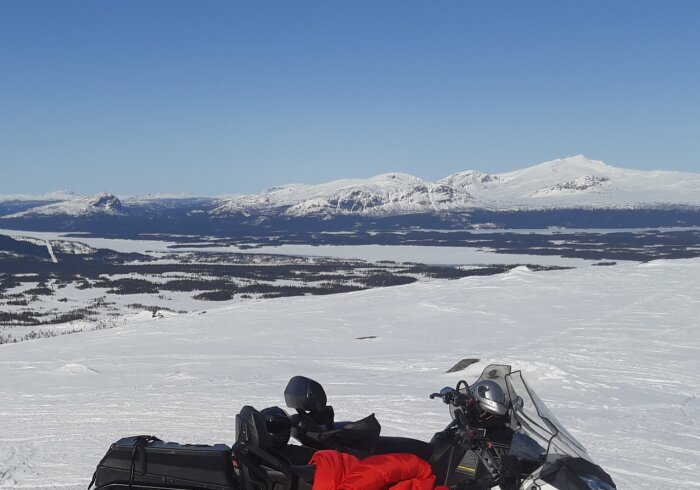 Två snöskotrar på snötäckt landskap, berg i bakgrunden, klar himmel, soligt och vintertid.