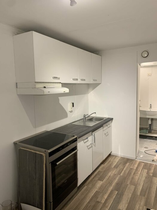 Ett minimalistiskt kök med vita skåp, grått golv, och en delvis synlig toalett i bakgrunden.