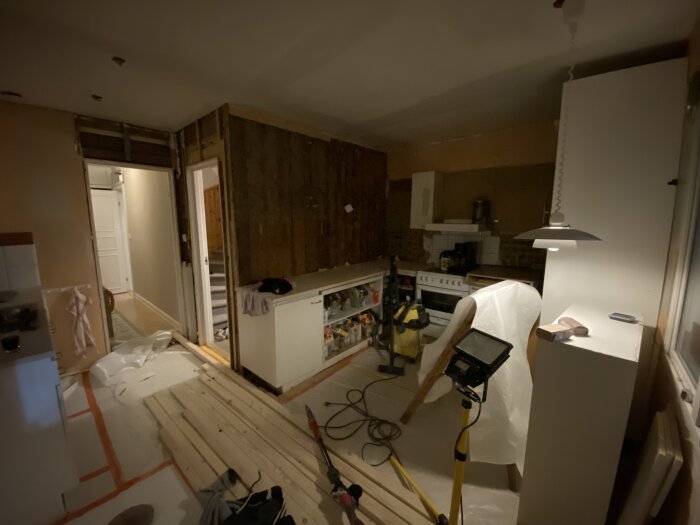 Ett kök under renovering, nakna väggar, byggmaterial, verktyg och delvis täckta möbler.