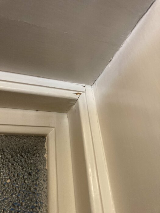 Vit dörrkarm och list med spricka där de möts, grå vägg, strukturerat glasfönster.