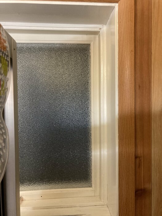 Frostat glasfönster i vit karm, träpanel intill, interiör vy, dämpat ljus, diskret plats, hemmiljö.