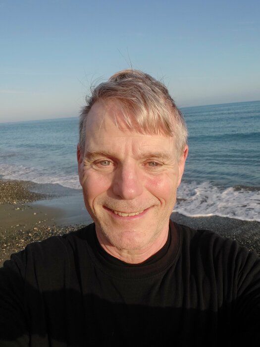 Man tar selfie framför havet, solsken, glad, strand, dagtid, avslappnad.