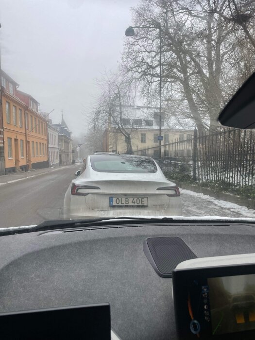Bild från bilens förarplats, disigt väder, stadsmiljö, gatuparkering, byggnader, nakna träd, europeisk registreringsskylt.