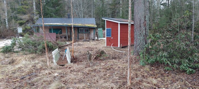 Enkel stuga och skjul i skogsmiljö, mossigt tak, solarpanel, avskalad natur, delvis snötäckt mark.