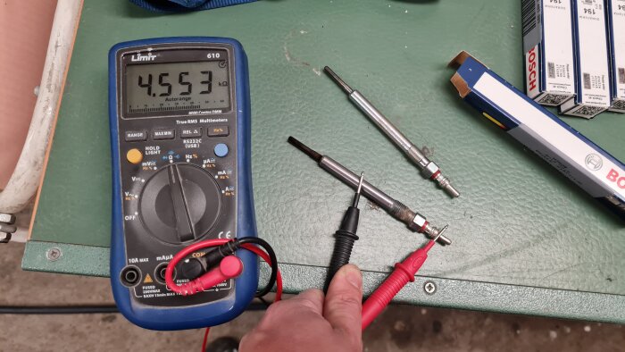 Multimeter visar resistans, hand håller prob, bänk med tändstift och förpackning.