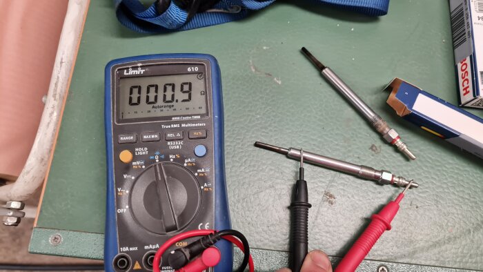 Multimeter, testledningar, glödstift och förpackning på verkstadsbänk.