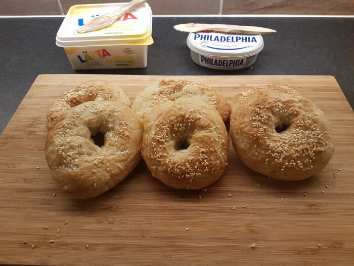 Fyra rostade bagels med sesamfrön, såstunnar och smör på skärbräda, redo för frukost.