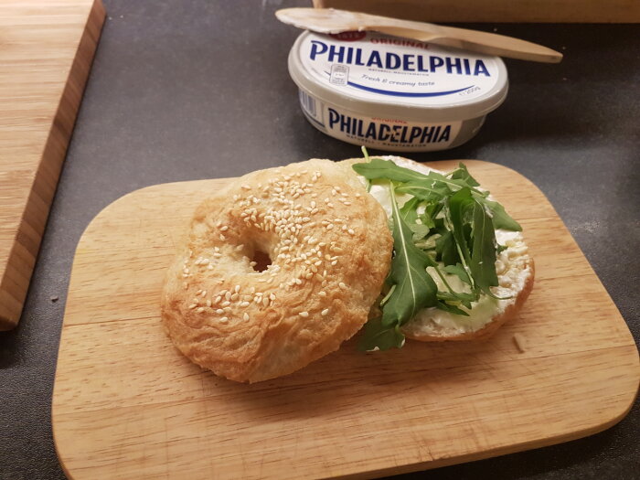 Sesam bagel med färskost och ruccola på träbräda, Philadelphia förpackning och kniv i bakgrunden.