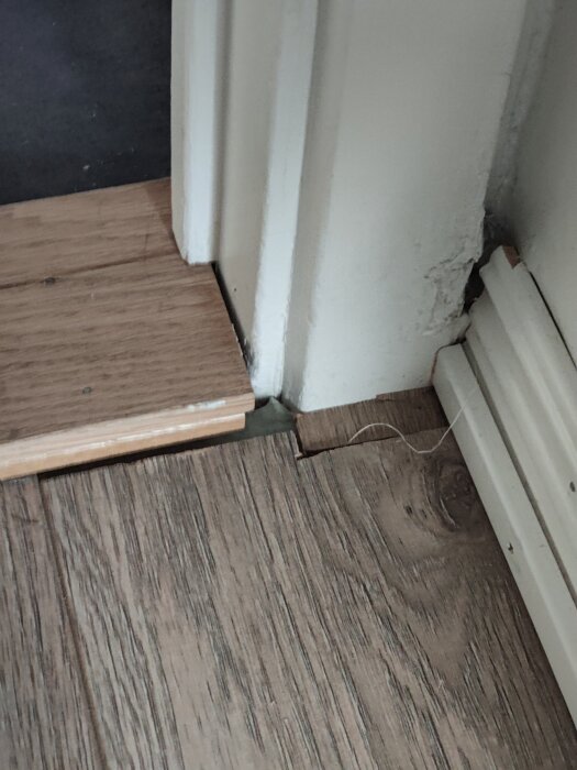 Skadad vägg vid golvlist, sliten färg, träimitationsgolv möter vit pelare och list. Reparationsbehov synligt.