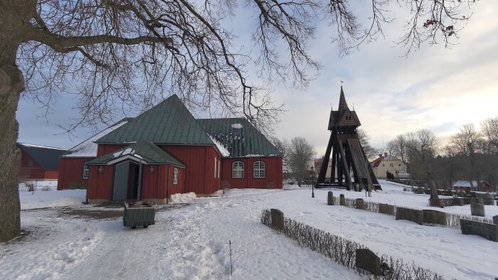 Vintrig kyrkogård med röda byggnader, klockstapel, snö, nakna träd, lugn atmosfär.