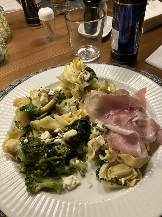 Pastatallrik med broccoli, fetaost och prosciutto på ett bord med dryck och kryddor.