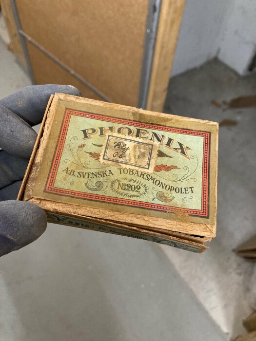 En hand i handske håller en gammal Phoenix tändsticksask från Svenska Tobaksmonopolet. Vintage, samlarobjekt, slitet, historiskt.