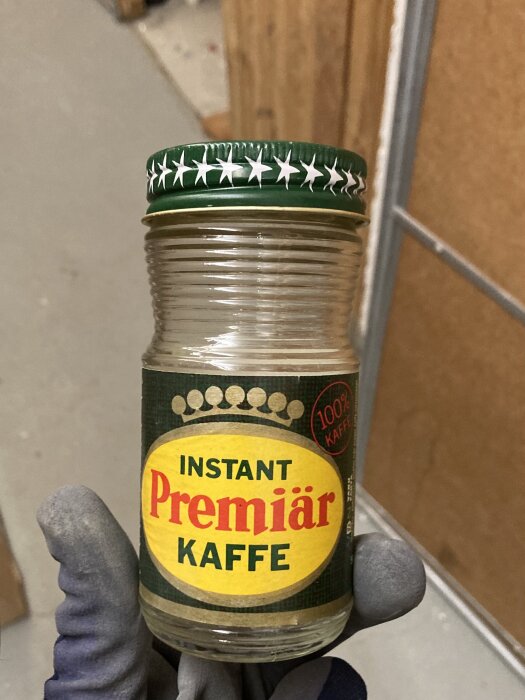 En person håller en burk med texten "INSTANT Premiär KAFFE" och "100% KAFFE", klädd i blå handskar, inomhus.