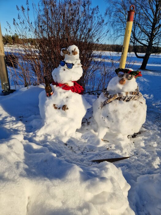 Två snögubbar med tillbehör står i snö, i soligt vinterlandskap, med buskar och klarblå himmel.