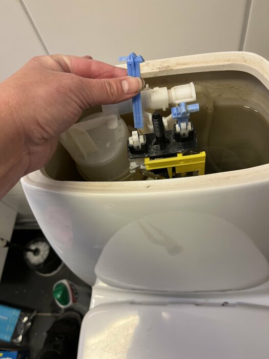 En hand justerar inuti en toalettstolens vattentank; rörmekanik synlig, underhåll eller reparation antyds.