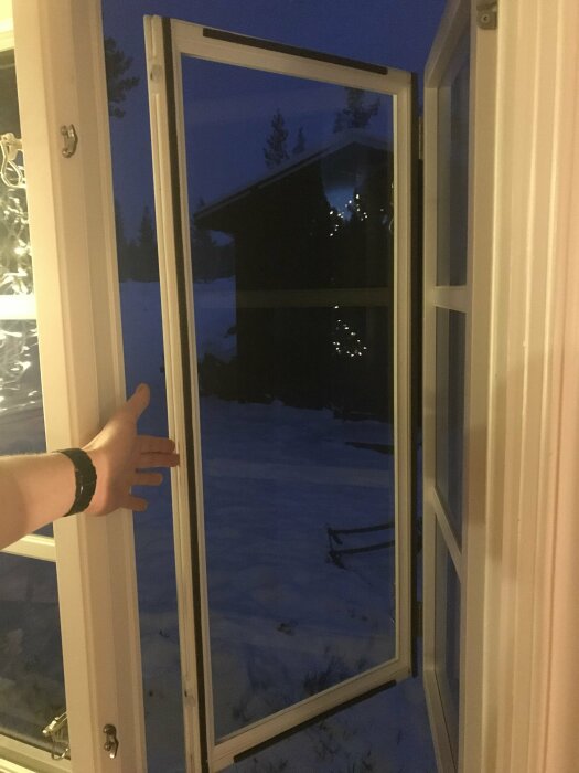 Hand öppnar fönster mot snöigt landskap med skymning och ljusdekorationer på byggnad.