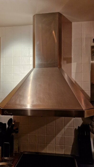 Köksfläkt i rostfritt stål över spis, vita kakelplattor i bakgrunden, köksredskap syns till höger.