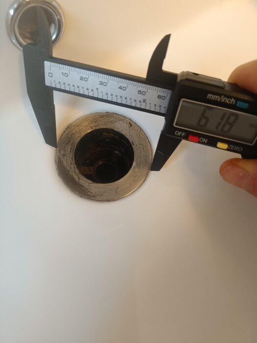 Digital skjutmått mäter diameter på vaskens avlopp. Indikerar cirka 61.8 mm. Vit bakgrund.