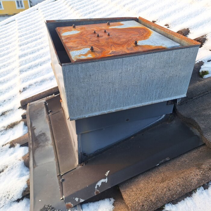 Ventilation på tak med snö, rostig lock, solbelyst, slitna ytor, bostadshus i bakgrunden.