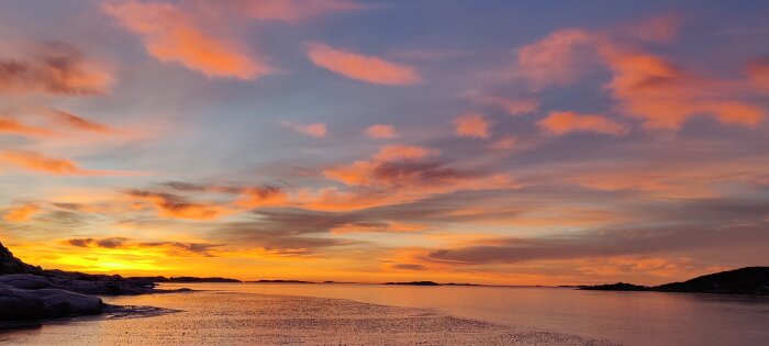 Solnedgång över hav, orangefärgade moln, klippor, vacker himmel, lugnt vatten, naturskön panorama.