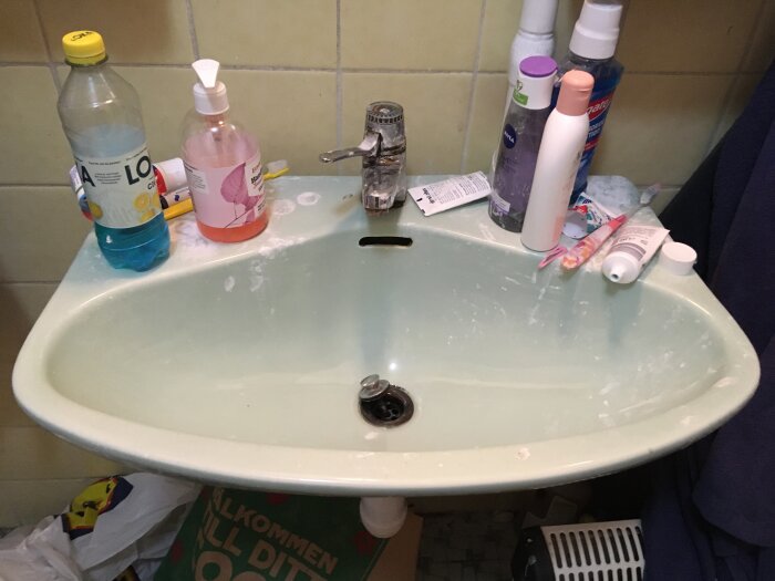 Ett rörigt badrums handfat med rengöringsprodukter, hygienartiklar och en tandborste.