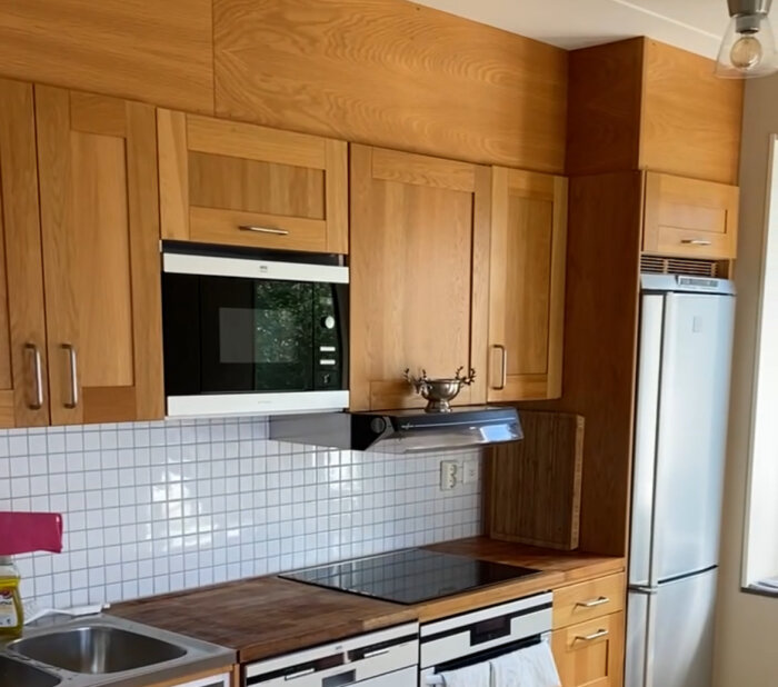 Ett kök med träskåp, vita kakelväggar, inbyggd mikro, spishäll, diskho och ett kylskåp.
