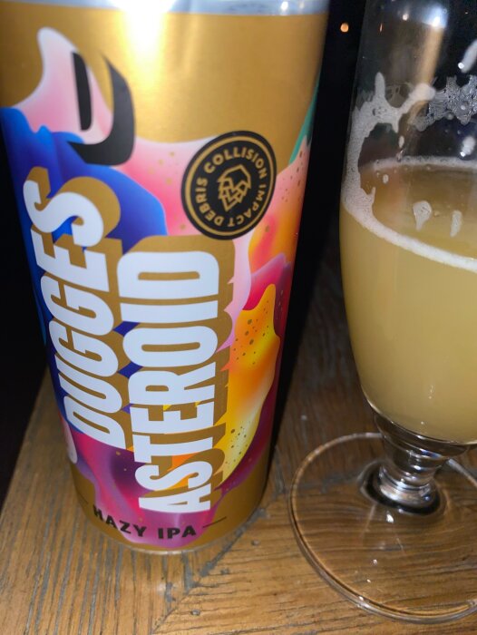 En färgglad ölburk märkt "Dugges ASTEROID Hazy IPA" bredvid ett halvfullt glas öl.