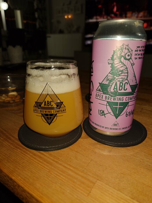 Ett glas öl med skum och en ölburk från Apex Brewing Company, placerade på träbord.